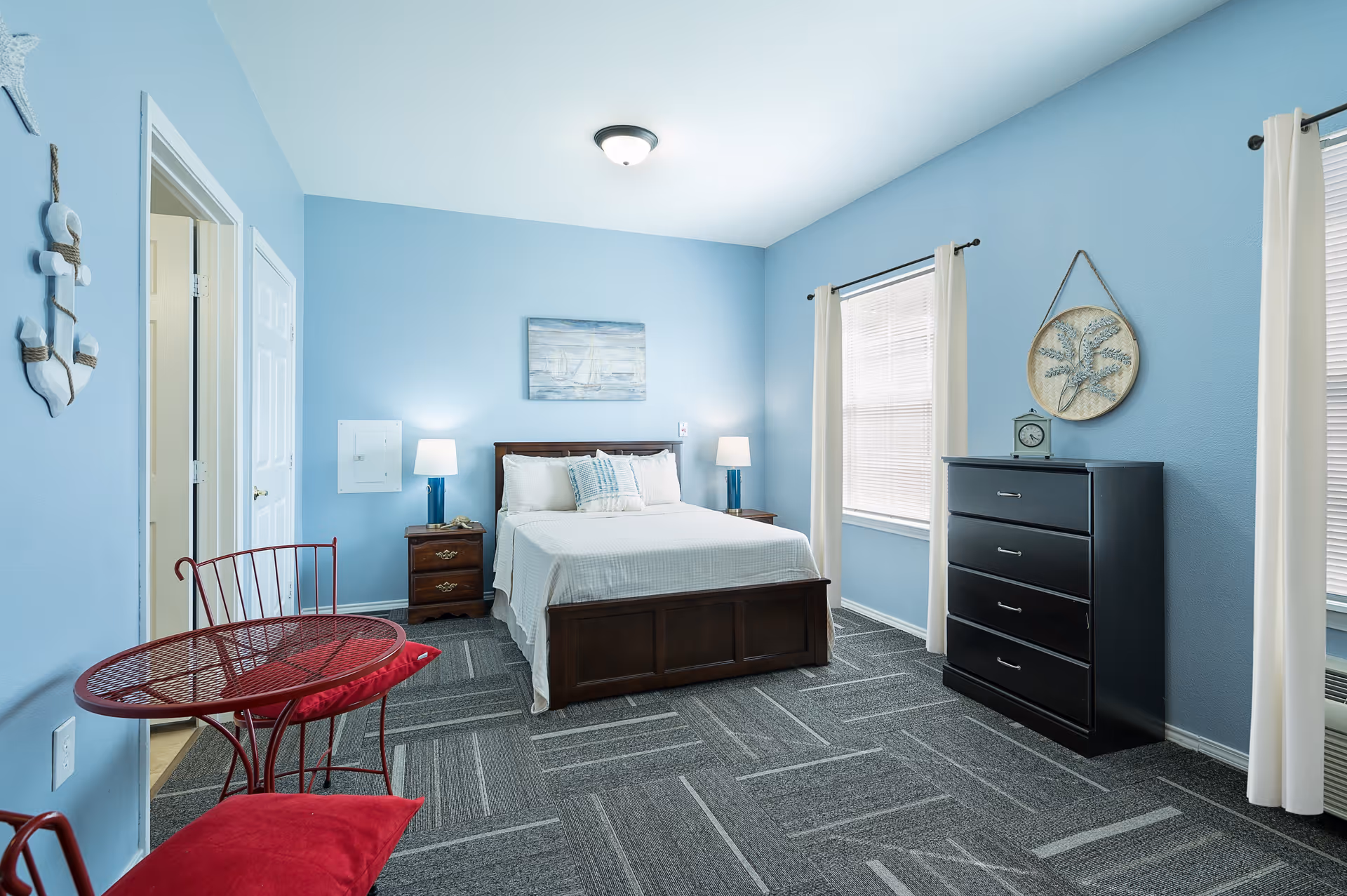 A bright bedroom with light blue walls, a double bed with white bedding and pillows, two bedside tables with lamps, a black chest of drawers, two windows with white curtains, a red metal table and chair set with red cushions, and nautical-themed wall decor including an anchor and a framed picture of a sailboat.