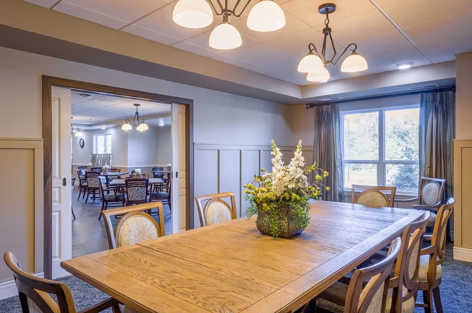 Well-lit dining room with a long wooden table, chairs, and a floral centerpiece, opening into another dining area.