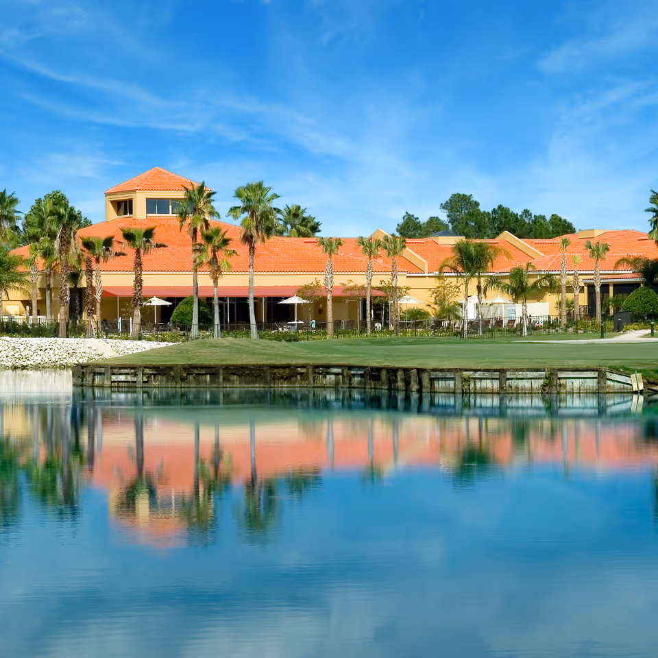 A large building with a red-tiled roof and beige walls is situated behind a calm body of water, reflecting the building and surrounding palm trees. The sky is clear and blue, and there is a well-maintained grassy area between the building and the water.
