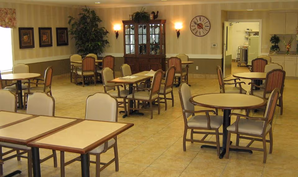 A dining room with multiple round and rectangular tables surrounded by chairs. The room has beige tiled floors, light-colored walls with a chair rail, framed artwork, a large clock, a wooden cabinet, and some plants. There is an open doorway leading to another room with kitchen equipment visible.