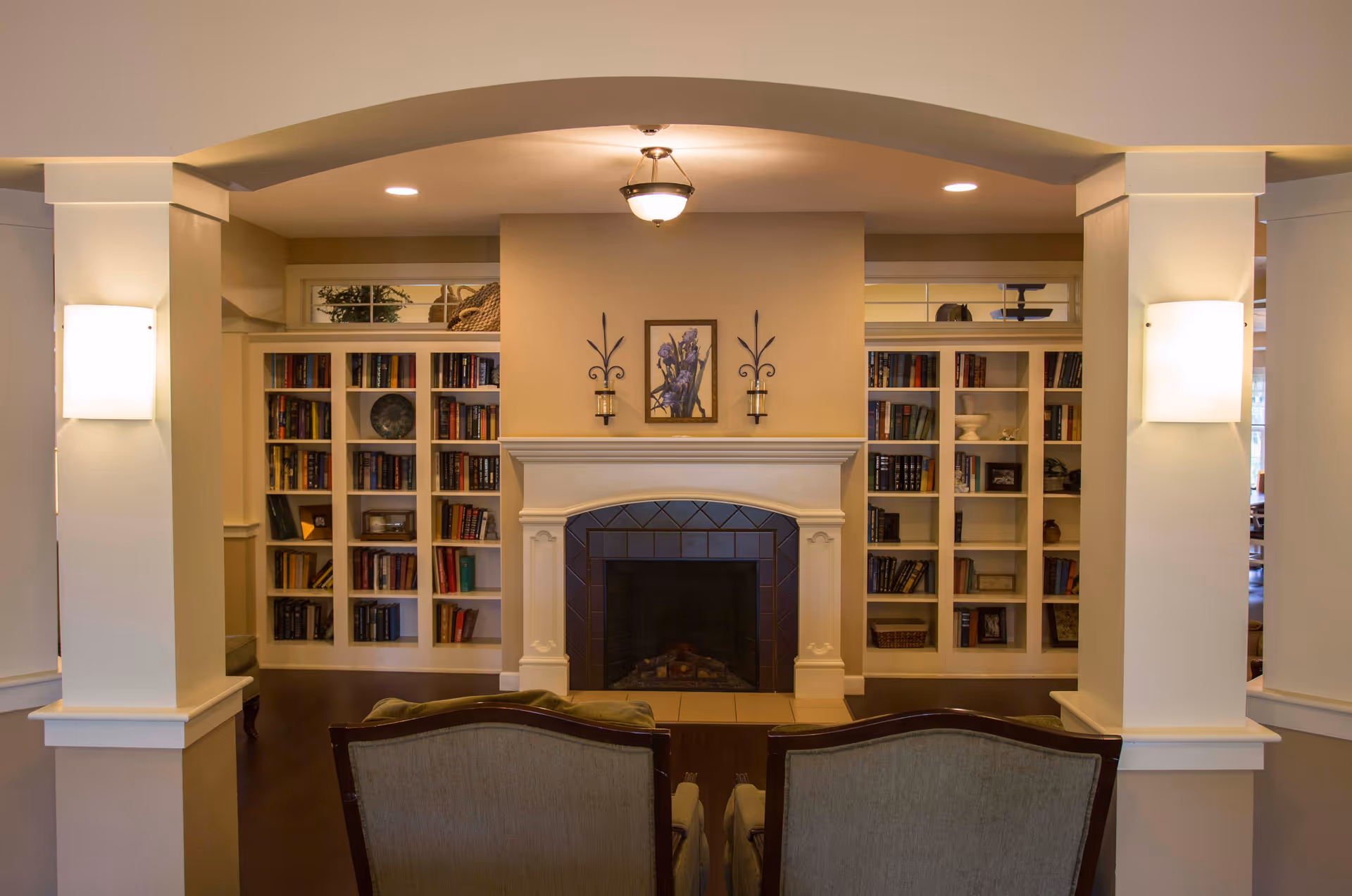 A cozy living room area with a central fireplace flanked by built-in bookshelves filled with books and decorative items. Two upholstered chairs face the fireplace. The room features warm lighting with wall sconces on either side of the fireplace and a ceiling light fixture above.