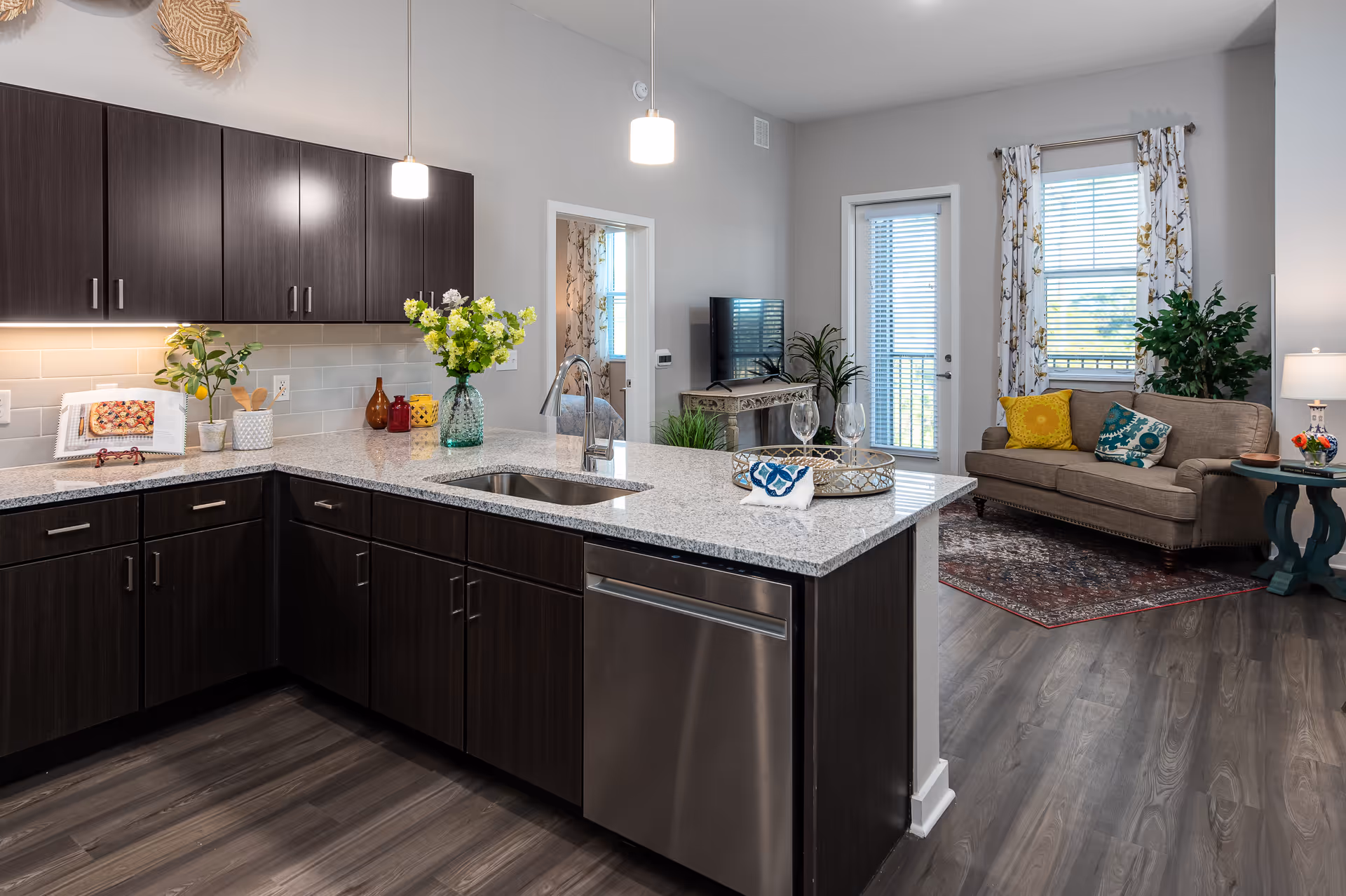 Modern kitchen with dark wood cabinets, granite countertops, and stainless steel dishwasher. The kitchen opens into a living room area with a beige sofa, colorful pillows, a patterned rug, a TV on a decorative console, and large windows with floral curtains. There are pendant lights hanging above the kitchen island and decorative plants throughout the space.