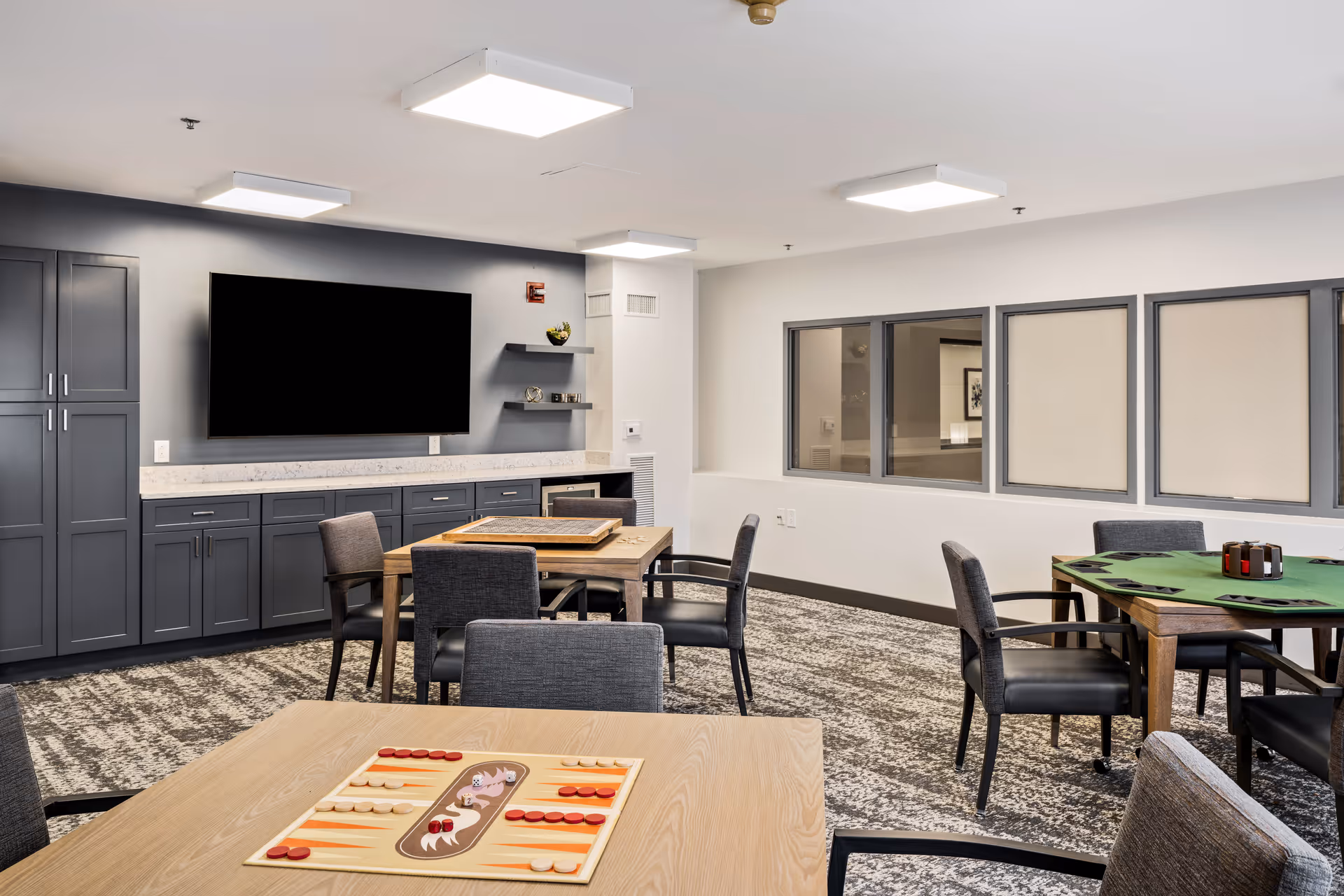 A senior living community common room with multiple tables and chairs arranged for games. One table in the foreground has a backgammon board set up. The room features gray cabinetry along one wall with a large flat-screen TV mounted above a countertop. The walls are light-colored, and there are windows on one side. The carpet has a patterned design, and the ceiling has modern square light fixtures.