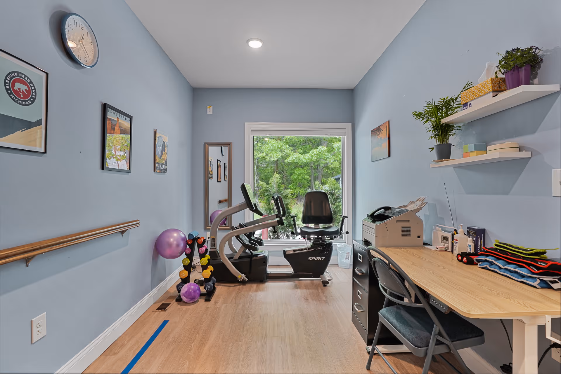 A small exercise room with light blue walls and wooden flooring. The room contains a recumbent exercise bike positioned in front of a large window showing green trees outside. On the left wall, there are framed pictures, a clock, and a wooden handrail. A rack with colorful dumbbells and exercise balls is placed near the bike. On the right side, there is a wooden desk with a chair, office supplies, a printer, and two white shelves holding plants and other items.