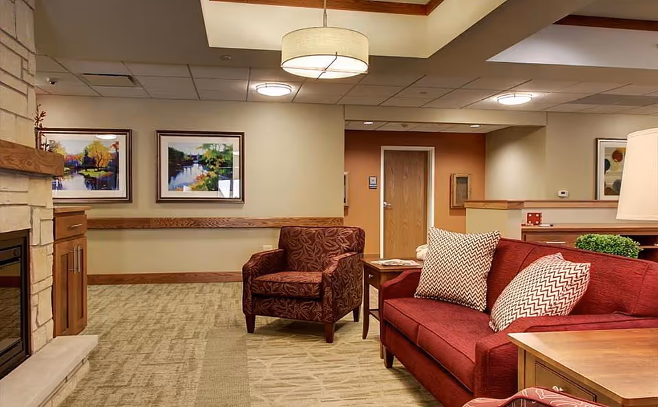 A cozy senior living facility common area with a red sofa adorned with patterned pillows, a matching armchair, wooden side tables, a stone fireplace on the left, and framed landscape paintings on the wall.