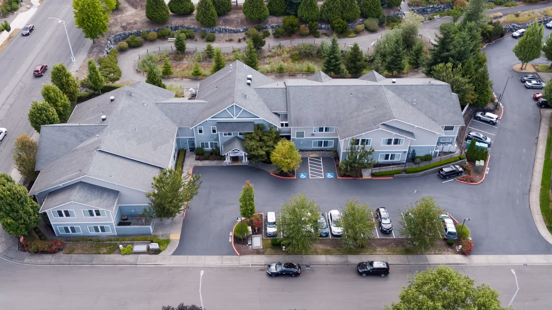 Aerial view of a senior living facility building with a gray roof and blue-gray exterior walls, surrounded by trees and parking spaces with several cars parked. The building is situated next to a road and landscaped areas with greenery.
