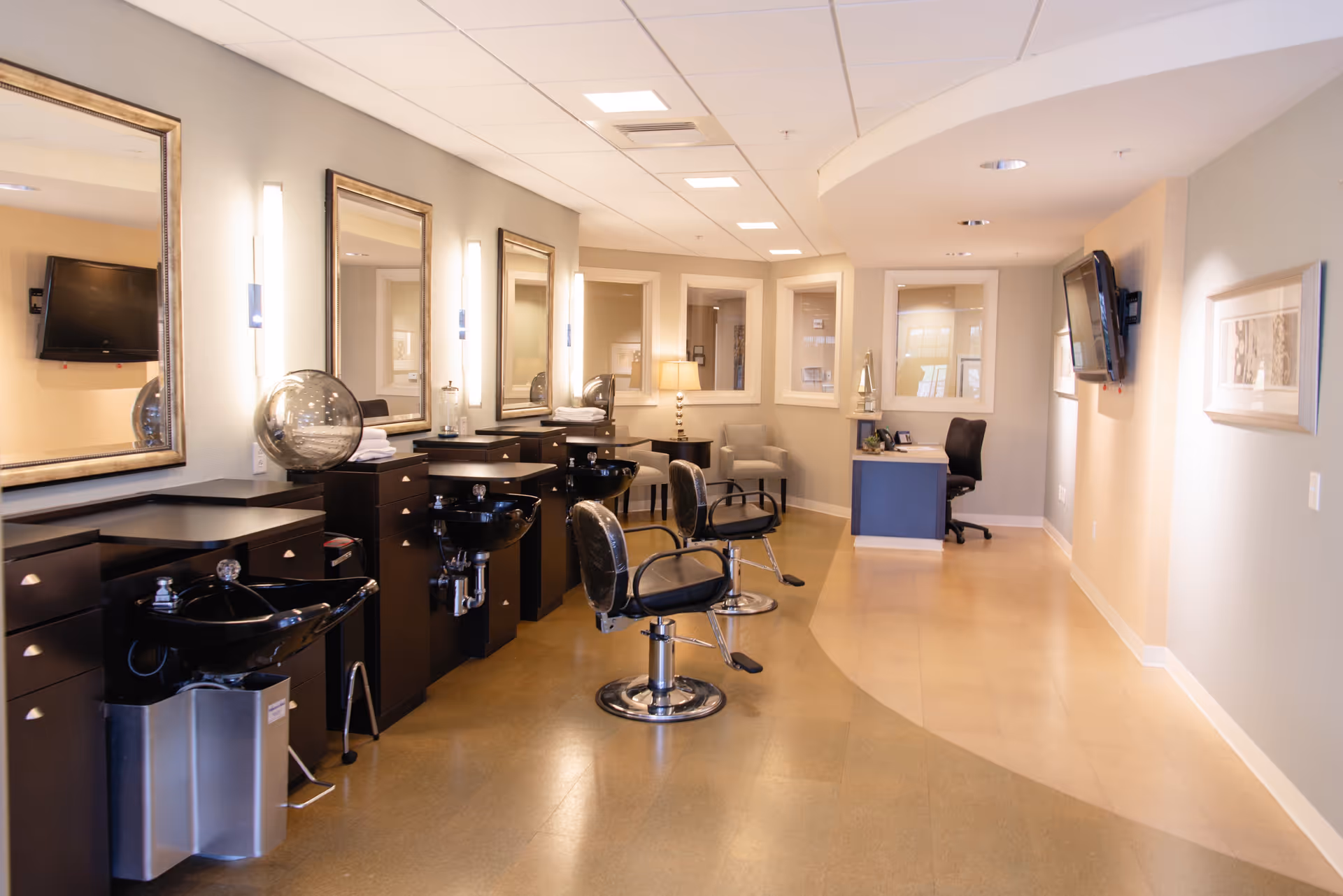 A well-lit salon area with three black salon chairs in front of three mirrors mounted on the wall. Each station has a black sink and dark cabinetry with drawers. The room has a light-colored floor and walls, with a desk and chair at the far end, a wall-mounted TV, and framed artwork on the walls.