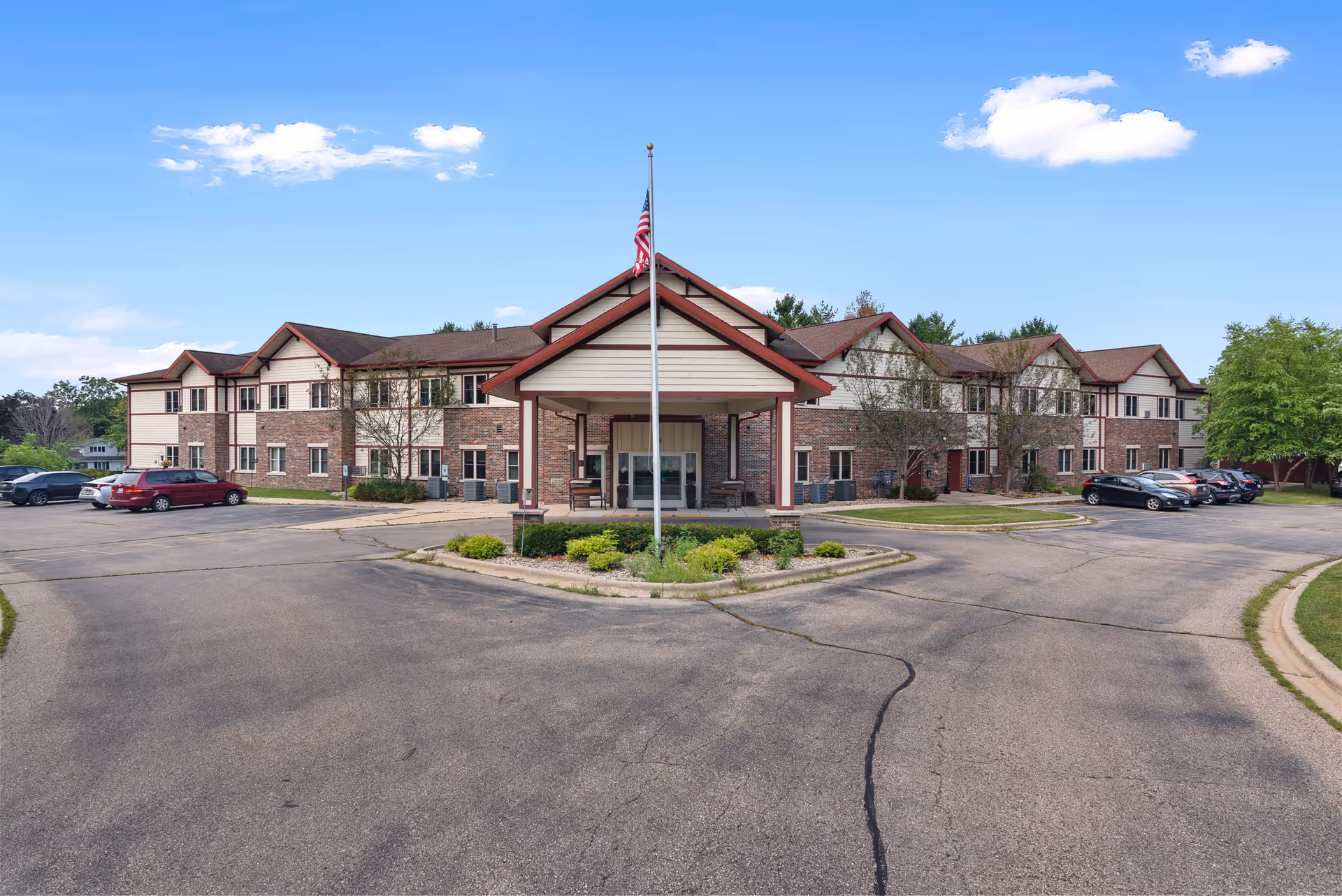 Exterior front view of a two-story senior living facility building with a covered entrance, an American flag on a flagpole, several parked cars, and a clear blue sky with a few clouds.