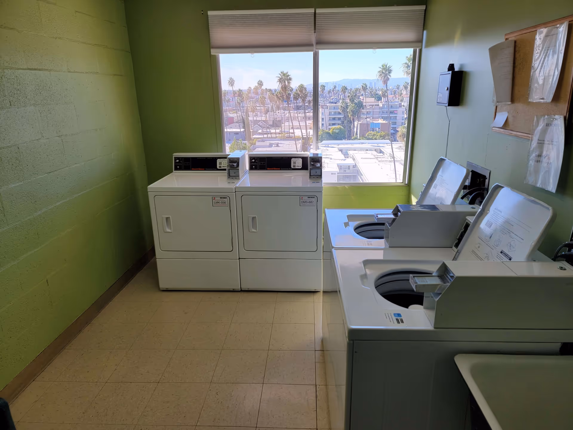 Laundry room with two white top-loading washing machines on the right and two white front-loading dryers on the left, a large window in the back showing a cityscape with palm trees and buildings, green painted walls, and a bulletin board on the right wall.