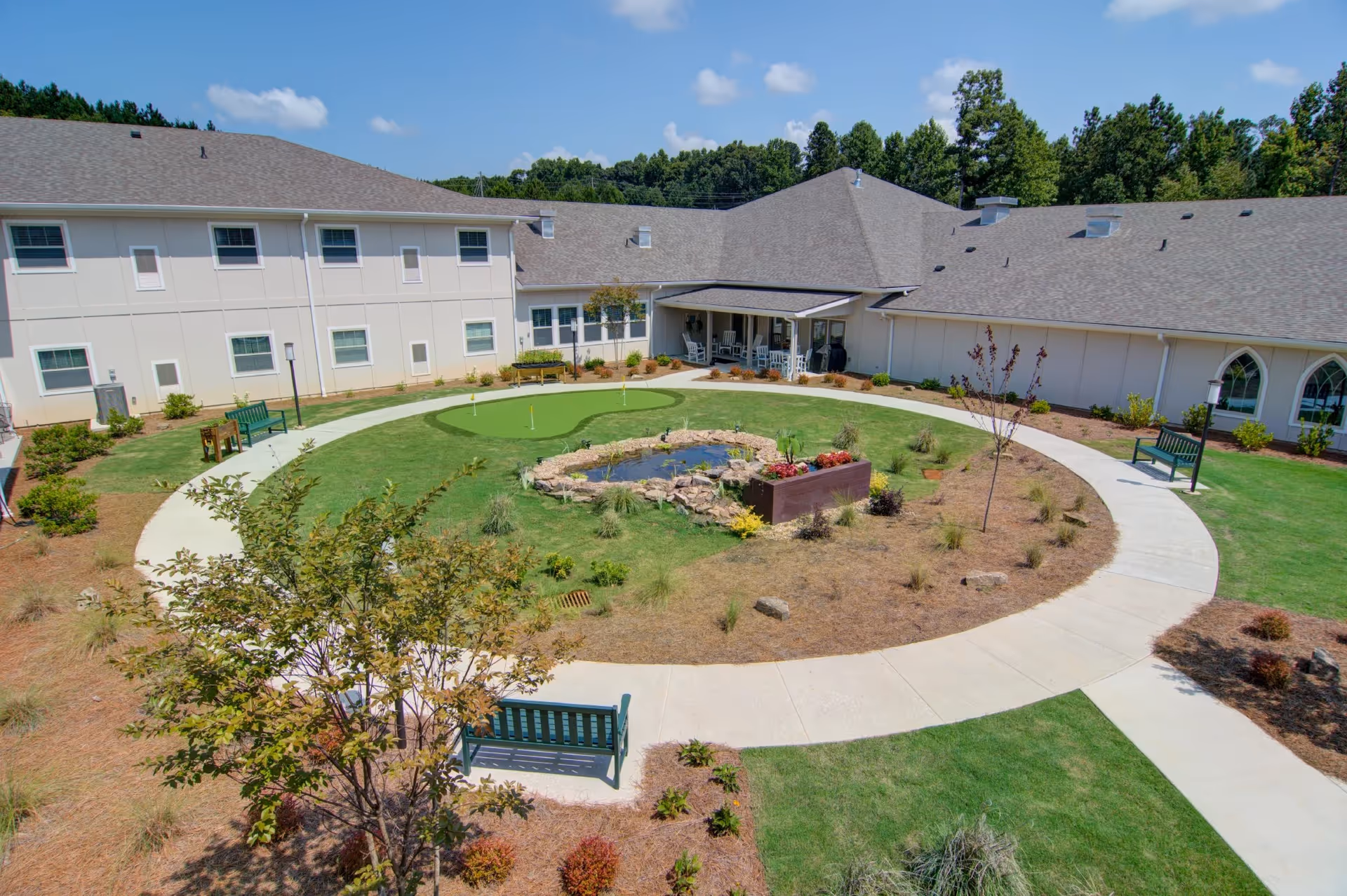 Outdoor courtyard area at Prema at Suwanee Creek featuring a circular walking path surrounding a small pond with rocks and plants, green benches, a putting green, and a covered porch with rocking chairs attached to a two-story building under a clear blue sky.