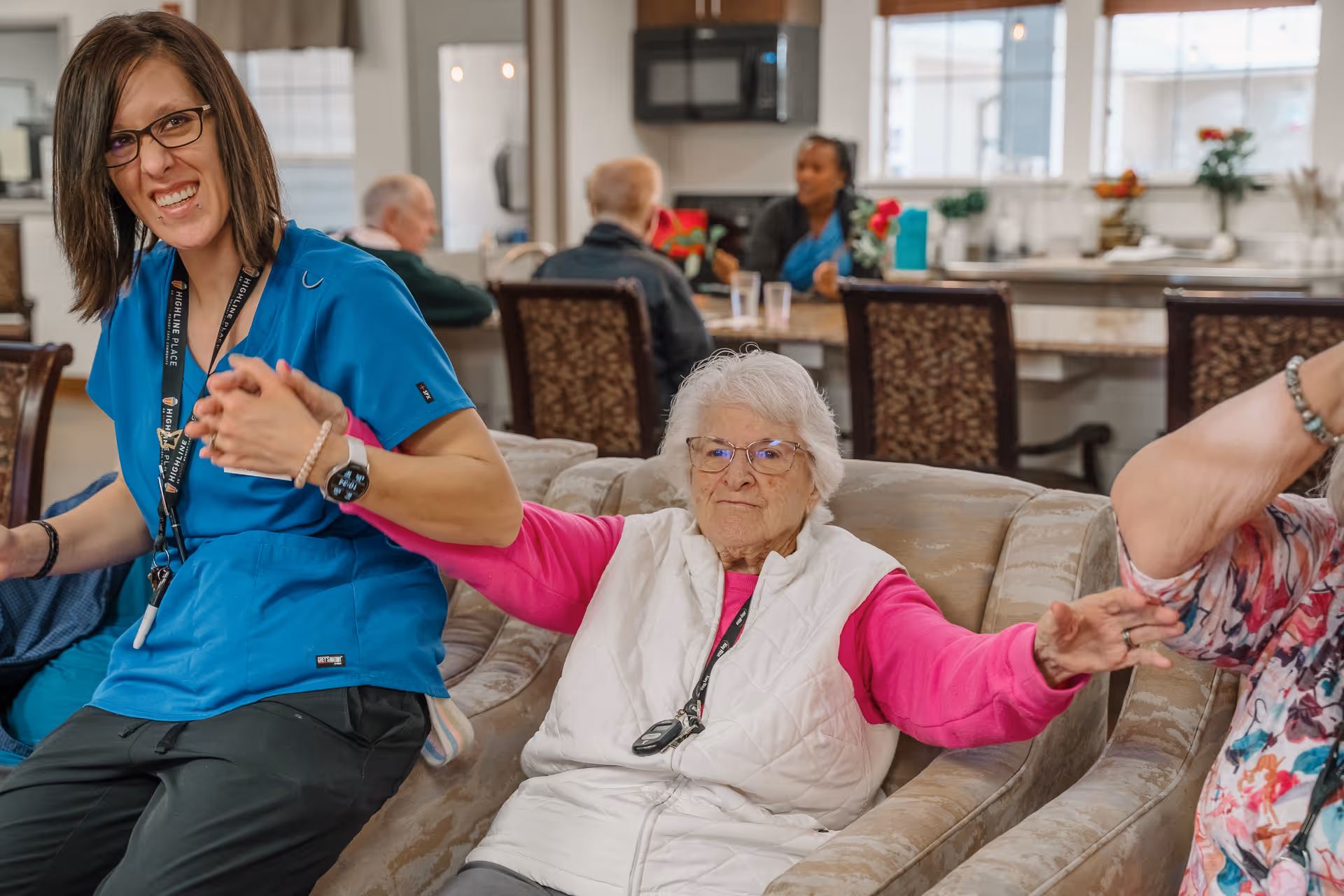 An elderly woman sits on a couch holding hands with caregivers in a communal living area with a kitchen and dining table in the background.