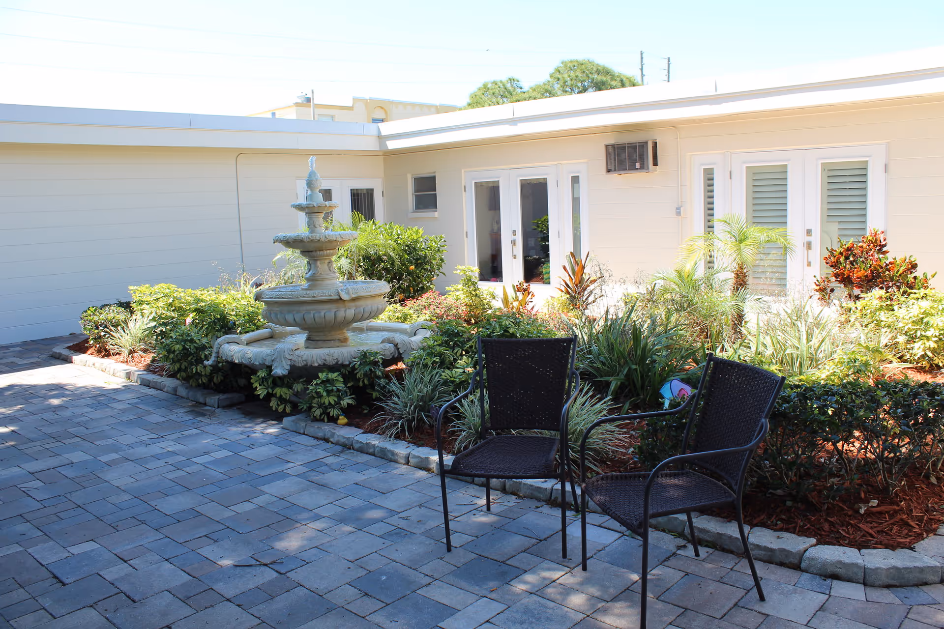 Outdoor courtyard area with a stone fountain surrounded by various green plants and shrubs. Two black metal chairs are placed on a paved stone patio in front of the fountain. The building exterior is light-colored with several doors and windows visible.