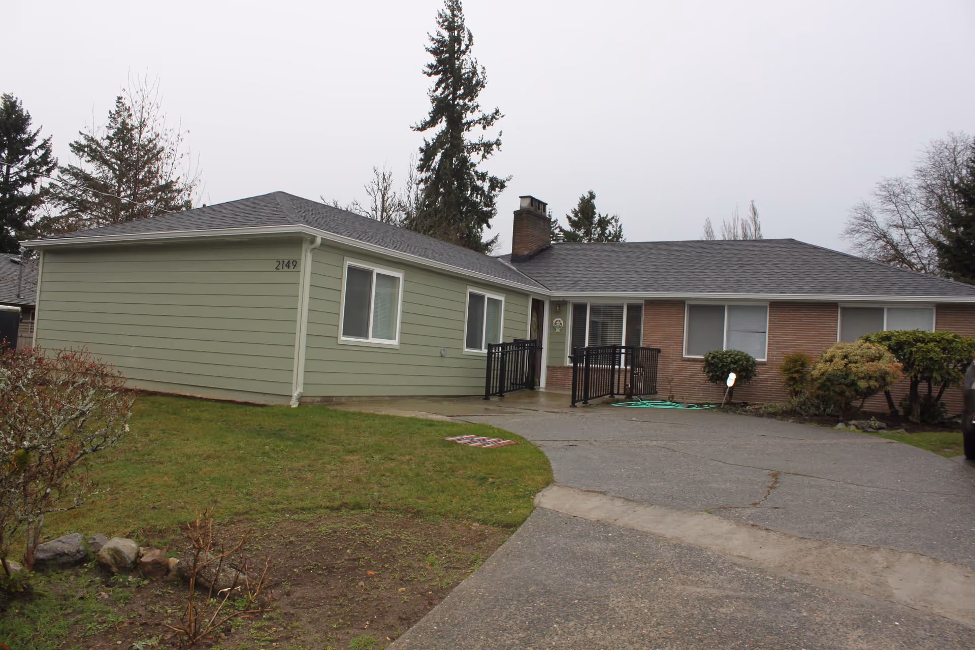 Exterior view of a single-story residential building with light green and brick siding, several windows, a black metal ramp leading to the front door, a driveway, and some bushes and trees around the property under an overcast sky.