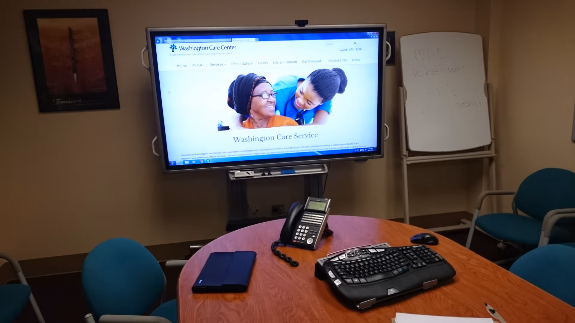 A small conference room with a round wooden table surrounded by teal office chairs. On the table are a telephone, a computer keyboard, a mouse, and a closed laptop or tablet. A large monitor on a stand displays the Washington Care Center website. In the background, there is a whiteboard with faint writing and a framed picture on the wall.
