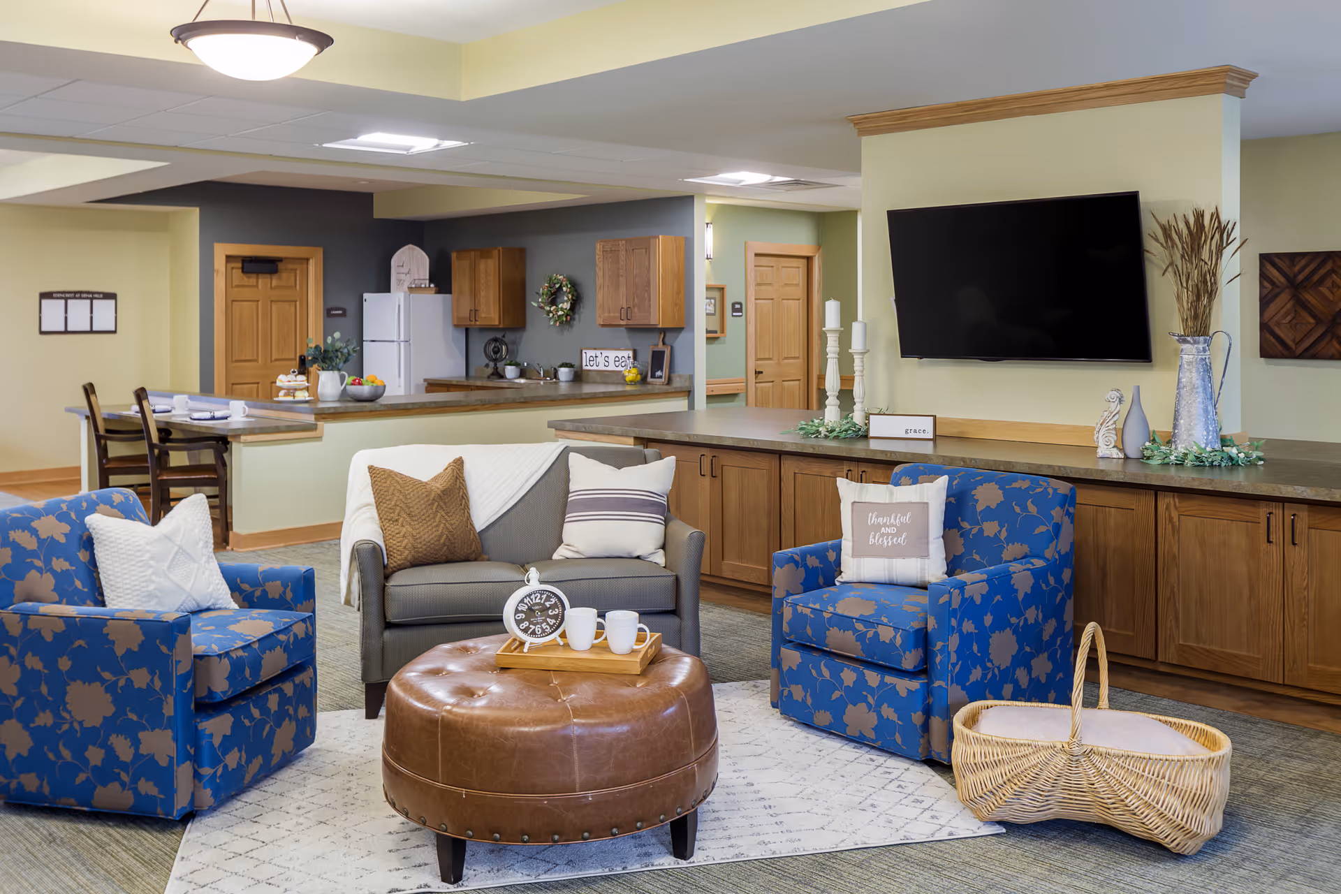 A cozy living room area in a senior living facility featuring two blue armchairs with floral patterns, a gray loveseat with pillows and a throw blanket, and a round brown leather ottoman with a tray holding two white mugs and a clock. Behind the seating area is a long counter with wooden cabinets, decorative items, and a large flat-screen TV mounted on a light green wall. In the background, there is a kitchen area with wooden cabinets, a white refrigerator, and a dining counter with chairs.