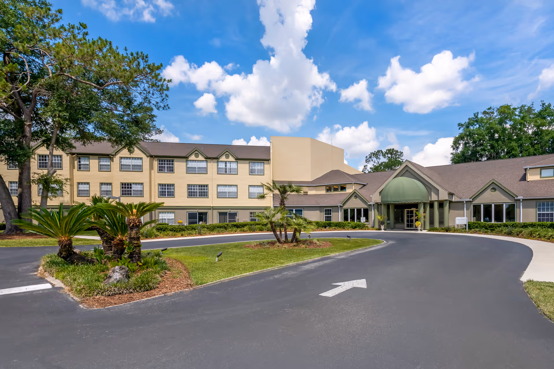 Exterior view of Brookdale Chambrel Pinecastle senior living facility showing a large building with multiple windows, a green awning over the entrance, a circular driveway with an arrow painted on the pavement, and landscaped greenery including palm trees and bushes under a partly cloudy blue sky.