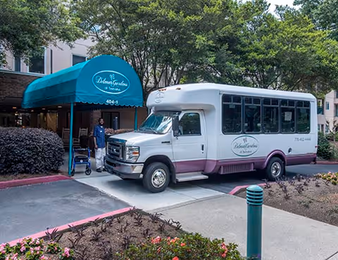 A white and purple shuttle bus parked in front of the entrance to Delmar Gardens of Smyrna, with a blue awning displaying the facility's name. A person in a blue uniform stands near the entrance holding a walker. The area is surrounded by trees and landscaping.