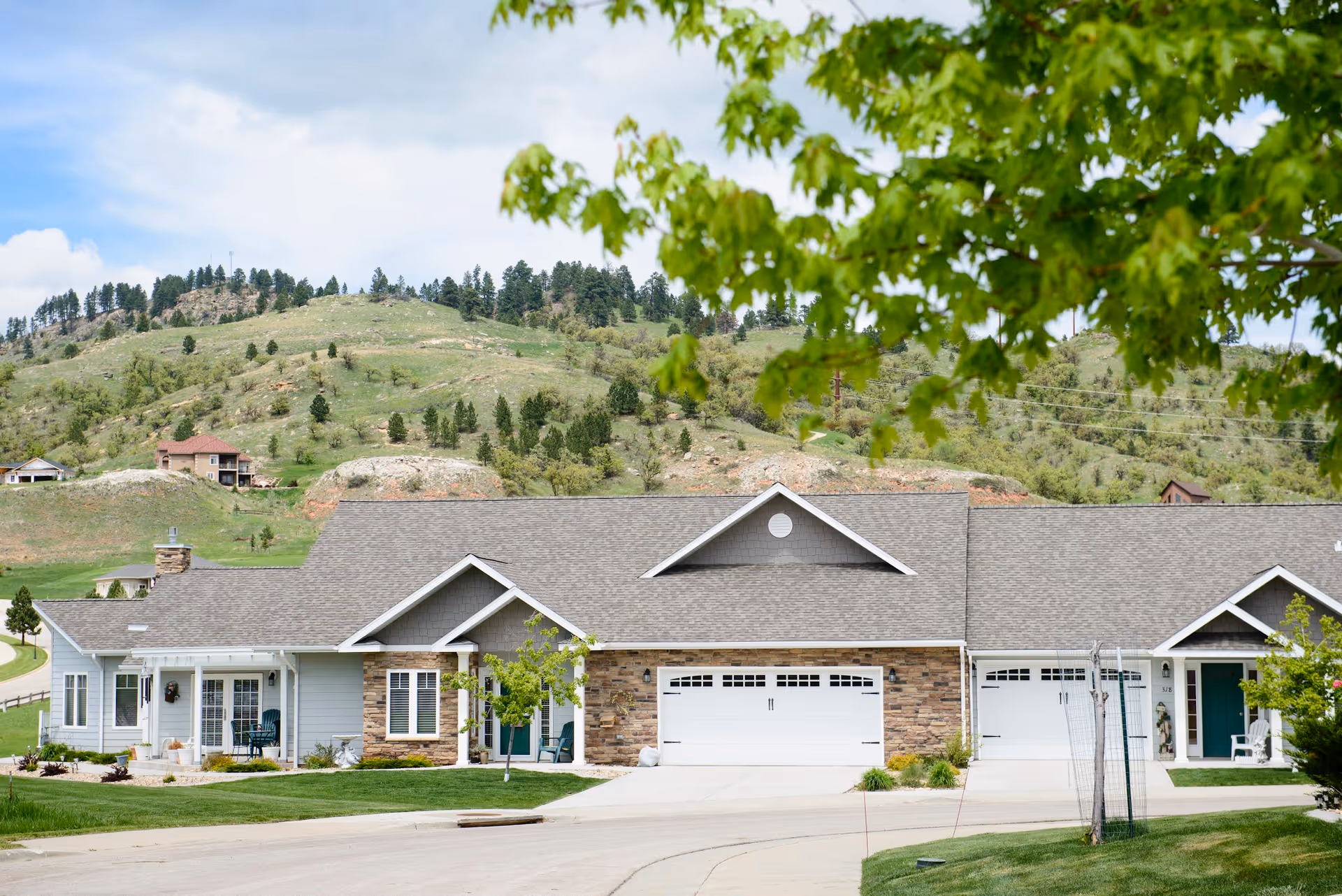 Single-story senior living building with attached garages, front porches, and landscaped lawn against a hillside backdrop.