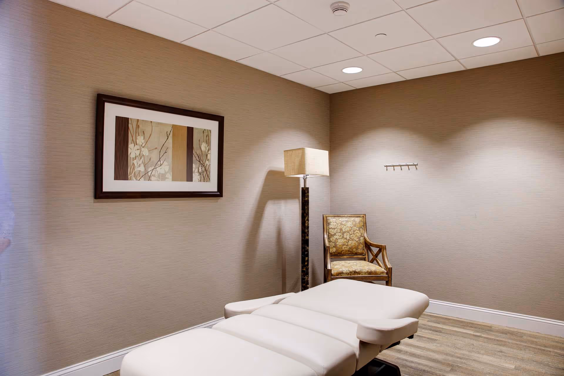 A small room with beige walls and wood flooring featuring a white padded massage or treatment table, a wooden chair with floral upholstery, a tall floor lamp with a beige shade, and a framed floral artwork on the wall.