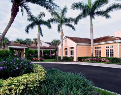 Exterior view of a senior living facility with peach-colored buildings, palm trees, and well-maintained landscaping including green shrubs and purple flowers under a partly cloudy sky.