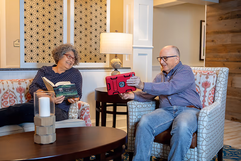 An elderly man and woman sitting in a cozy living room area. The woman is holding a book and smiling, while the man is holding a red tablet and showing it to her. They are seated on patterned armchairs with a round wooden coffee table in front of them, which has a decorative candle. A table lamp and decorative wall panels are visible in the background.