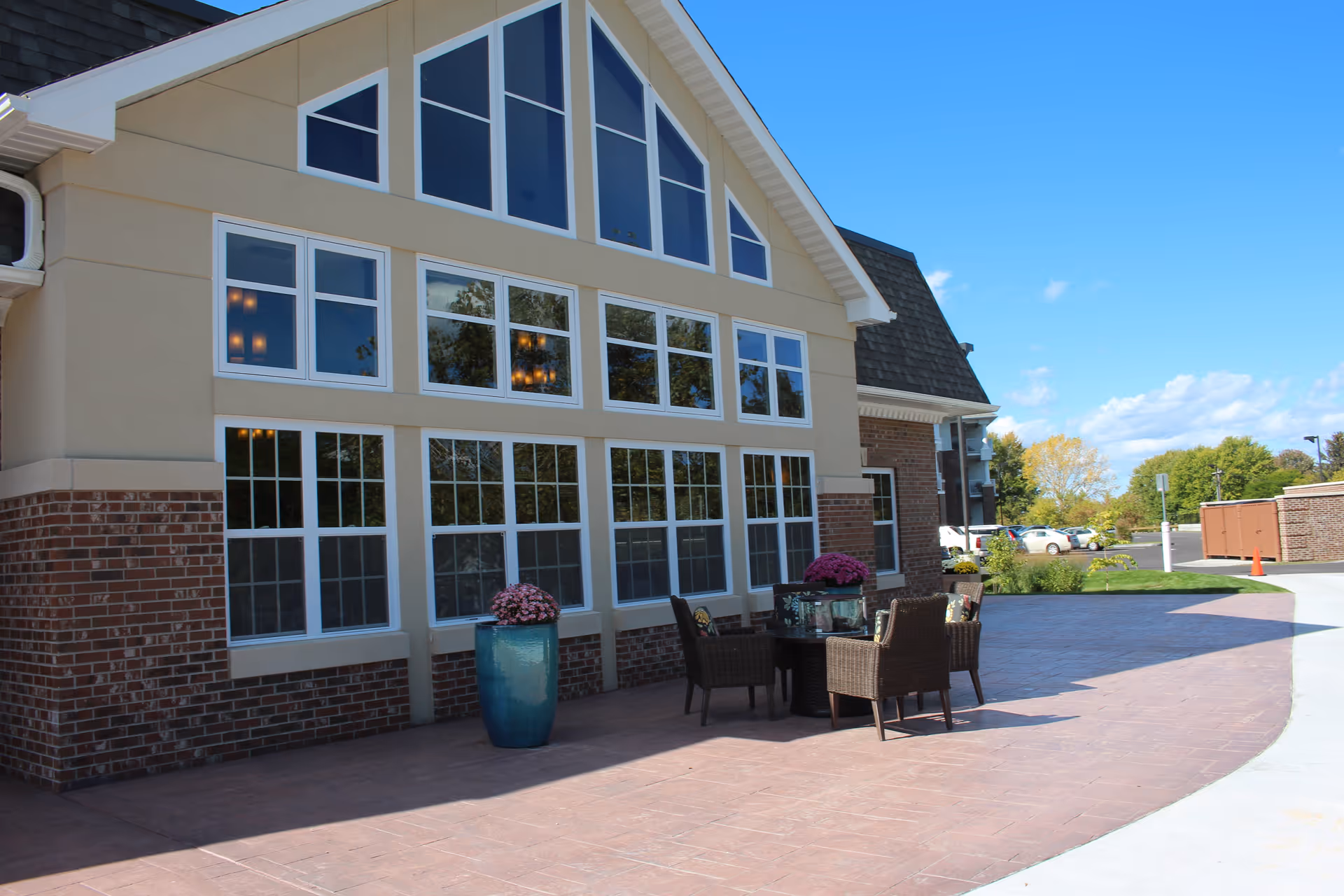 Outdoor patio area of a building with large windows, a table with four wicker chairs, and two large blue planters with pink flowers. The sky is clear and blue with some trees and parked cars in the background.