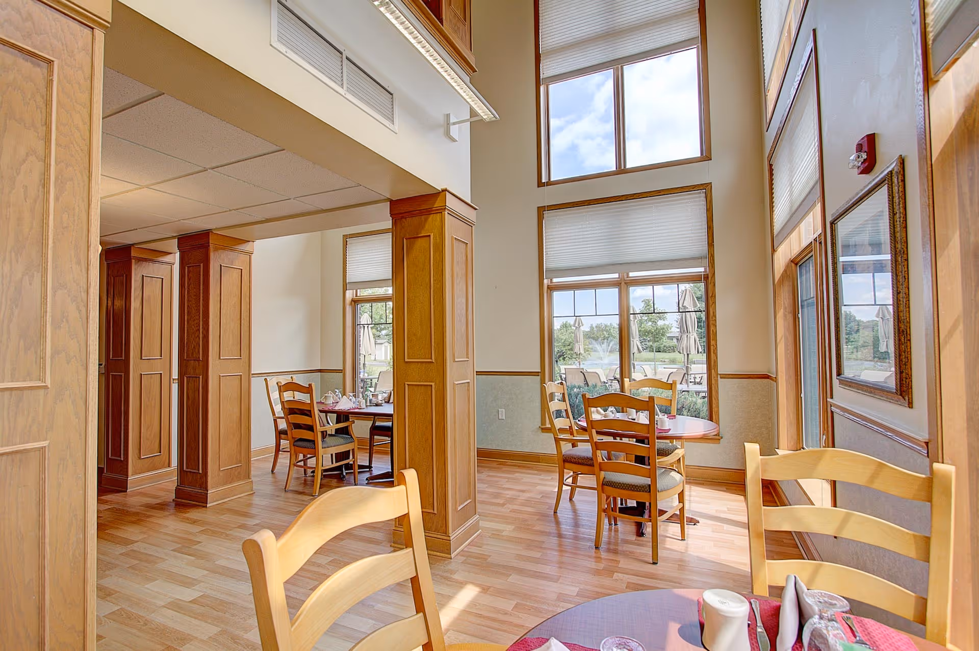 Sunlit dining room with wooden tables and chairs and tall windows overlooking an outdoor patio and fountain.