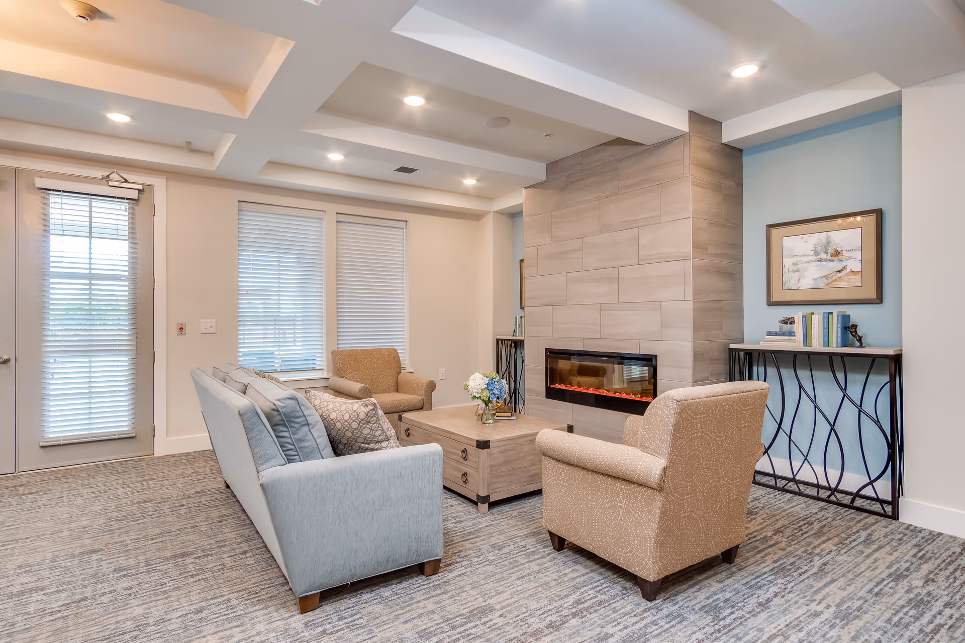 A cozy living room area with a light blue sofa, two beige armchairs, a wooden coffee table with a flower vase, and a modern electric fireplace set in a tiled wall. There is a console table with books and a framed painting on the wall behind one armchair. The room has large windows with blinds and recessed ceiling lights.