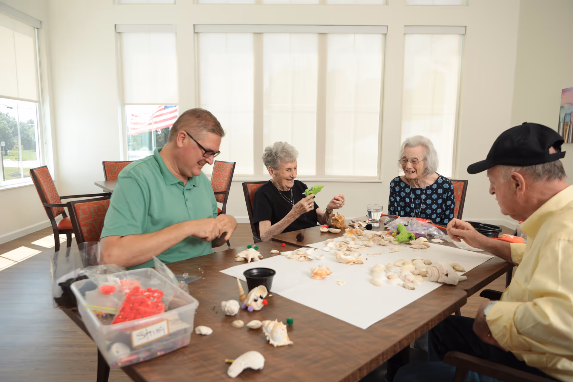 Four people sitting around a table in a bright room with large windows, engaging in a craft activity involving seashells and small decorative items. The group includes two elderly women, one elderly man wearing a black cap, and a younger man in a green polo shirt. The table is covered with seashells, craft supplies, and a white sheet of paper. An American flag is visible outside the window.