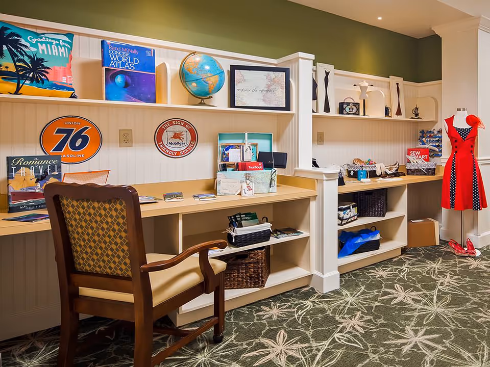 Well-lit interior activity/craft room with a long work counter and shelves filled with books, decor and supplies, a wooden chair, and a red dress on a mannequin.