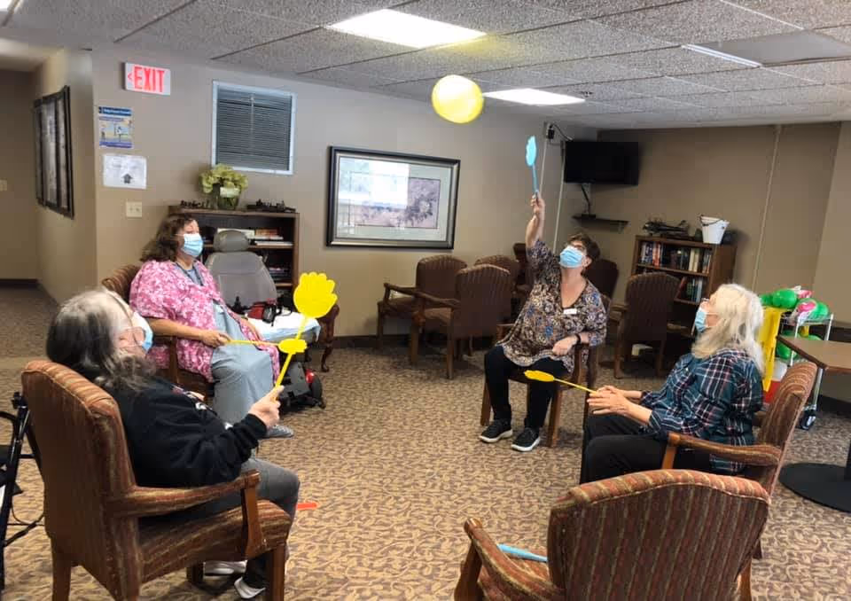 Four elderly women and a caregiver wearing face masks are seated in a circle in a common room, playing a game with yellow paddles and a yellow balloon. The room has carpeted floors, beige walls, framed artwork, and several chairs arranged around the space.