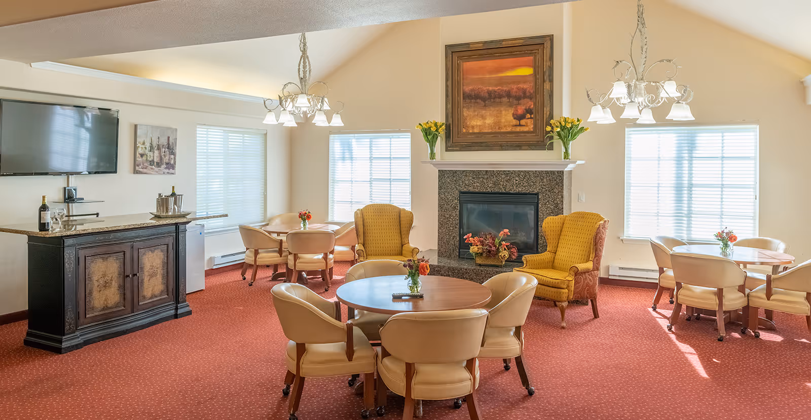 A cozy living room area with red carpet, beige walls, and multiple seating arrangements including round tables with beige chairs and two yellow armchairs near a fireplace. The fireplace has a granite surround and a framed painting above it. There are two chandeliers hanging from the ceiling, windows with blinds letting in natural light, a flat-screen TV mounted on the wall, and a cabinet with wine bottles and glasses on top.