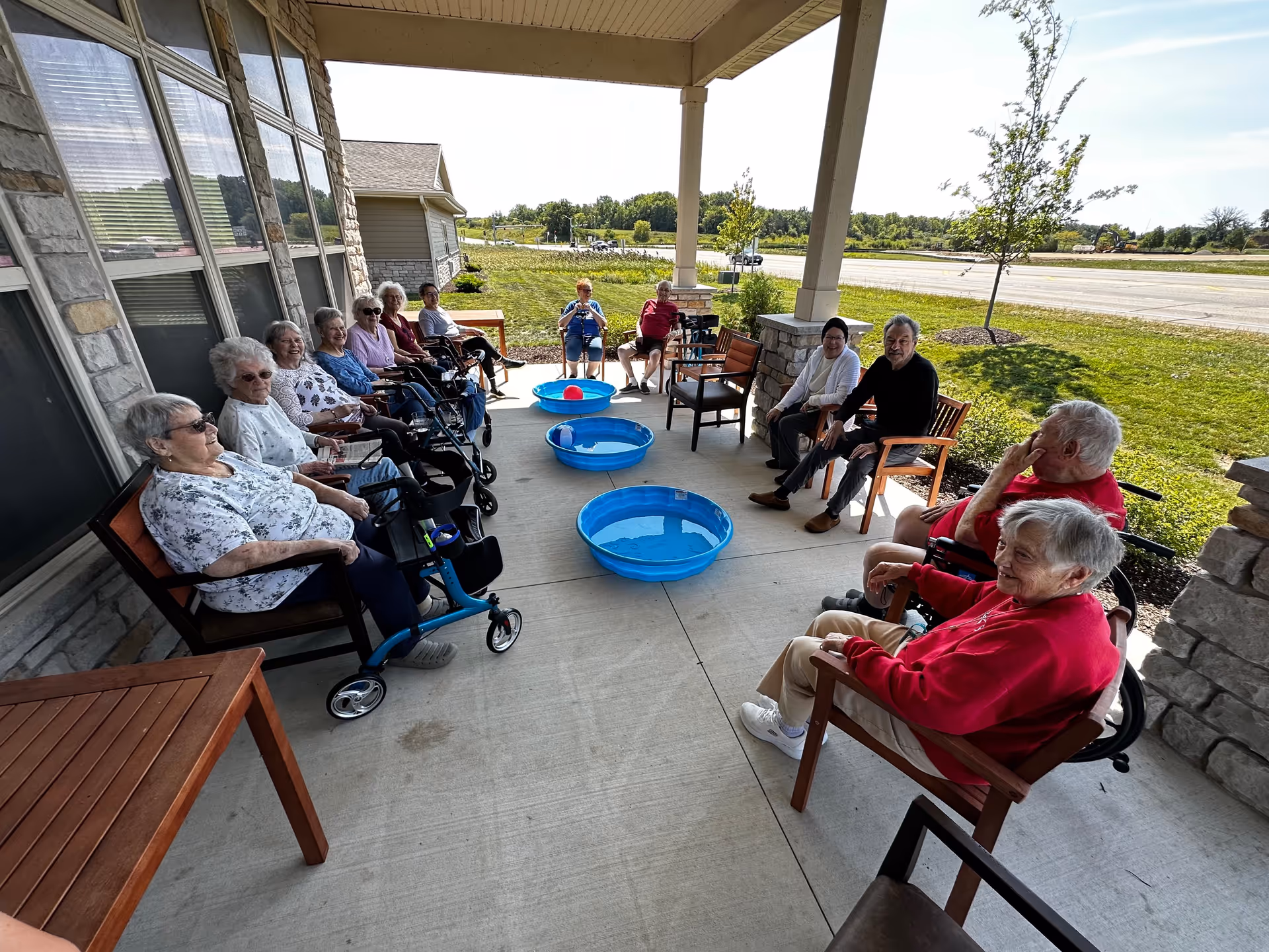 A group of elderly people sitting in chairs arranged in a semi-circle on a covered patio outside a building. There are three small blue plastic pools filled with water placed on the ground in the center of the group. The background shows a grassy area and a road with some trees and vehicles in the distance.