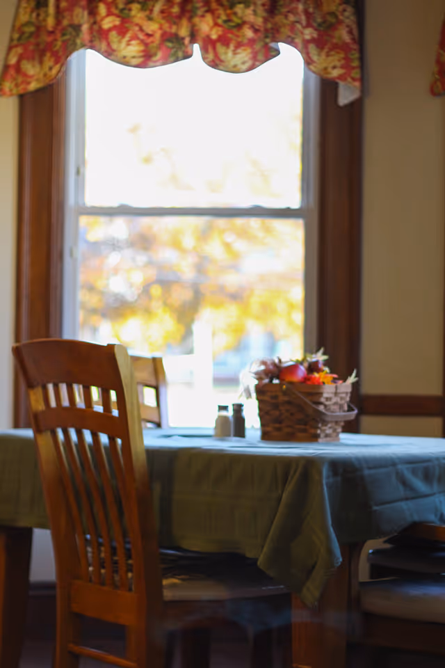 Sunlit dining table with wooden chairs, a green tablecloth, and a wicker centerpiece by a window.