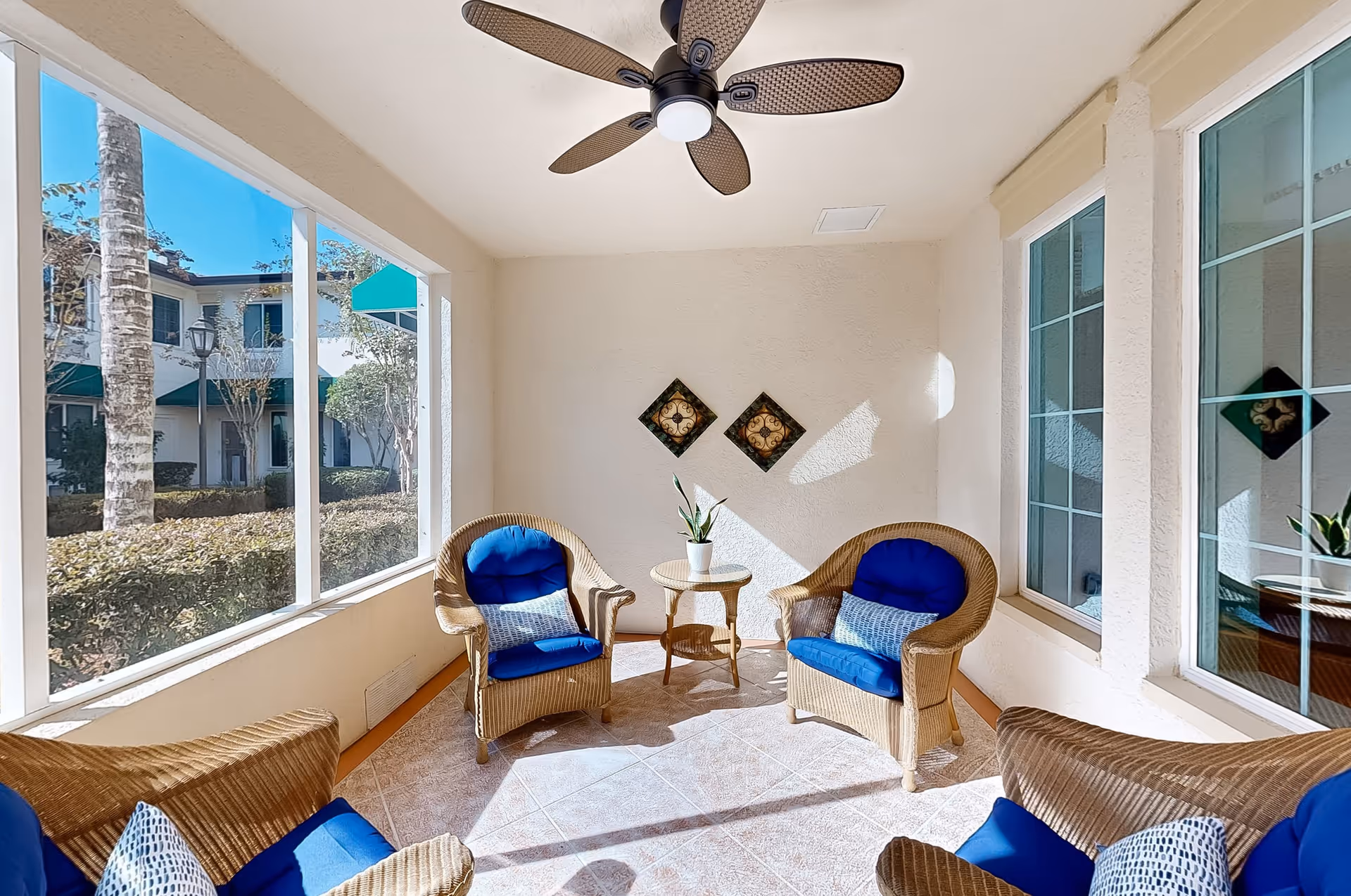 A bright sunroom with wicker chairs featuring blue cushions and patterned pillows, a small round wicker table with a potted plant, a ceiling fan with woven blades, and large windows showing an outdoor view of trees and a building with green awnings.