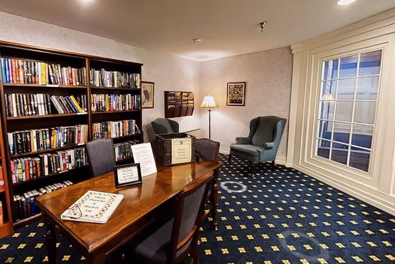 A cozy library room with a wooden table and four chairs in the center. The table has signs and a basket on it. Behind the table is a large bookshelf filled with books. To the right, there are two comfortable armchairs near a floor lamp and a window with white trim. The room has patterned carpet and soft lighting.