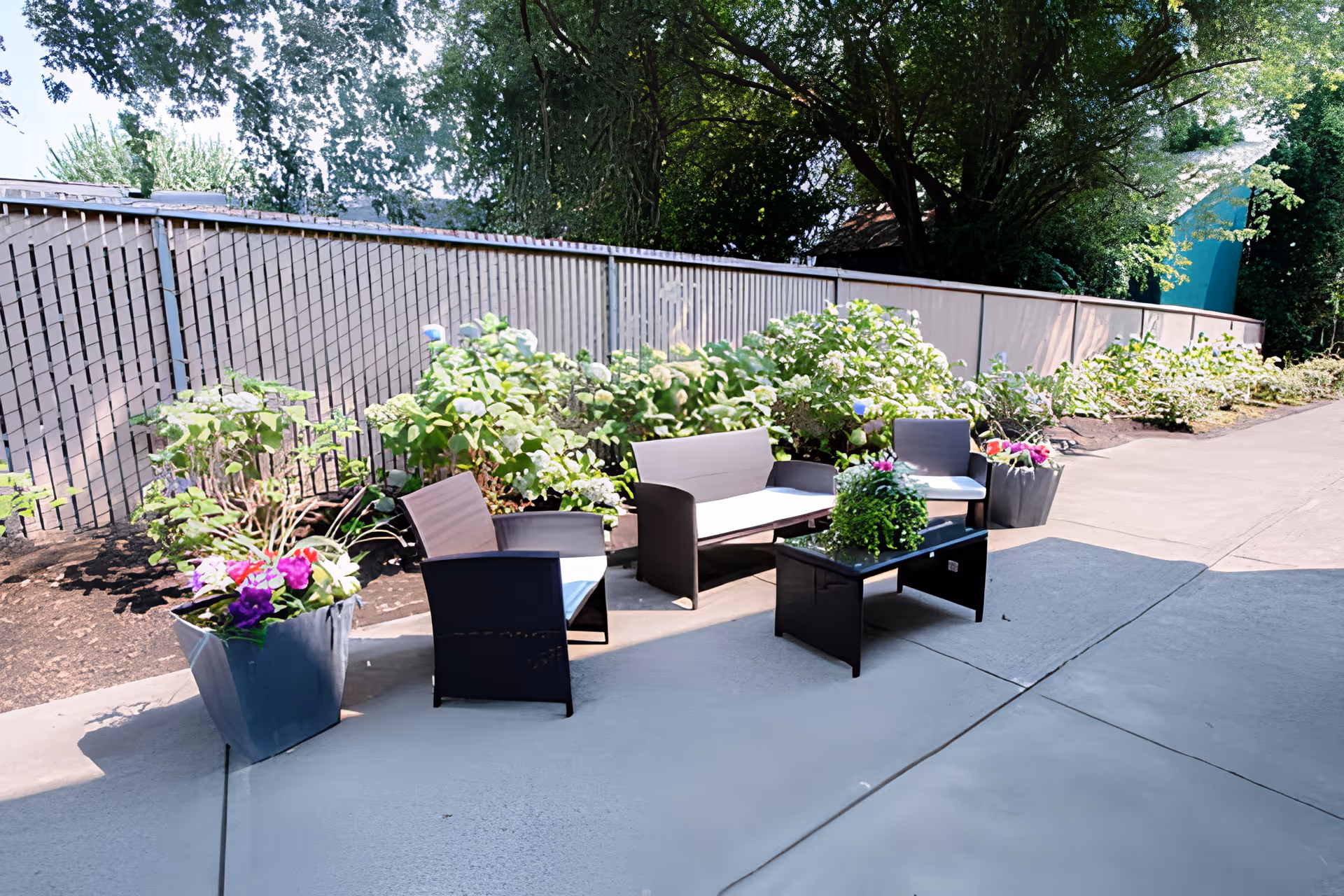 Outdoor patio area with modern black wicker seating including two chairs, a loveseat, and a glass-top coffee table, surrounded by large planters with colorful flowers and green shrubs along a wooden fence under shade from trees.