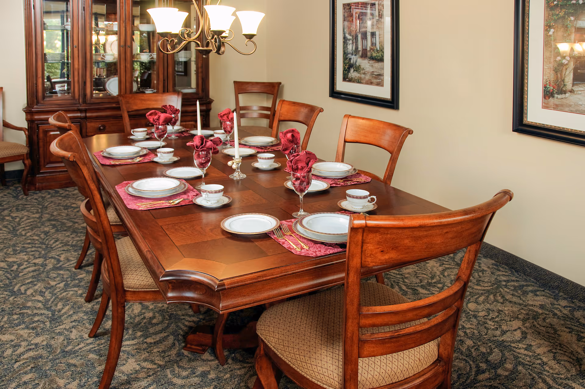 A formal dining room with a wooden dining table set for six people. The table has white plates, teacups, wine glasses with folded red napkins, and two white candles in glass holders. There are six wooden chairs with cushioned seats around the table. A wooden china cabinet with glass doors is visible in the background, along with framed artwork on the beige walls and a chandelier overhead.