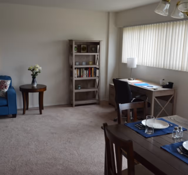 A cozy living area with beige carpet, a blue armchair with a floral cushion, a small round wooden side table with a vase of white flowers, a wooden bookshelf with books and decorative items, a wooden desk with a black office chair and a lamp, and a dining table set with plates, glasses, and salt and pepper shakers near a window with vertical blinds.