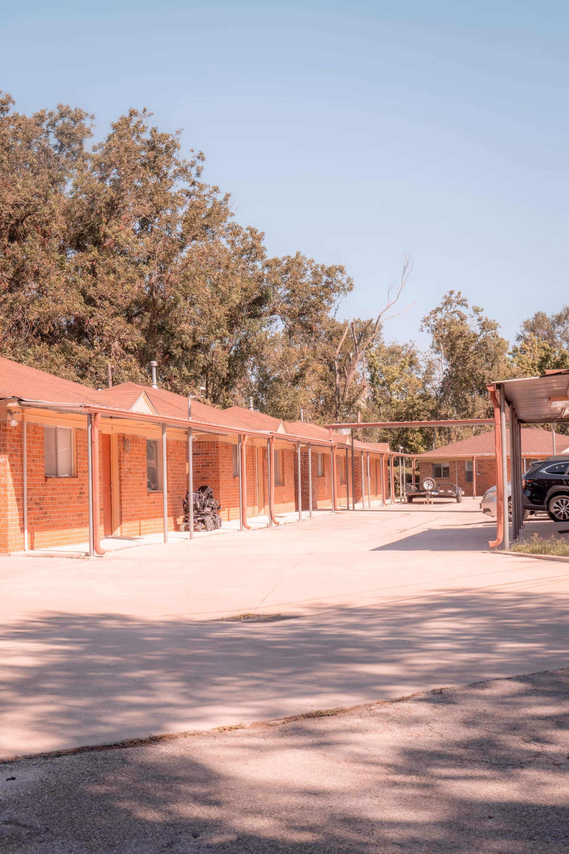 Exterior view of a single-story brick building with multiple doors and windows, covered walkways supported by metal poles, and a parking area with cars and a trailer. Trees are visible in the background under a clear sky.