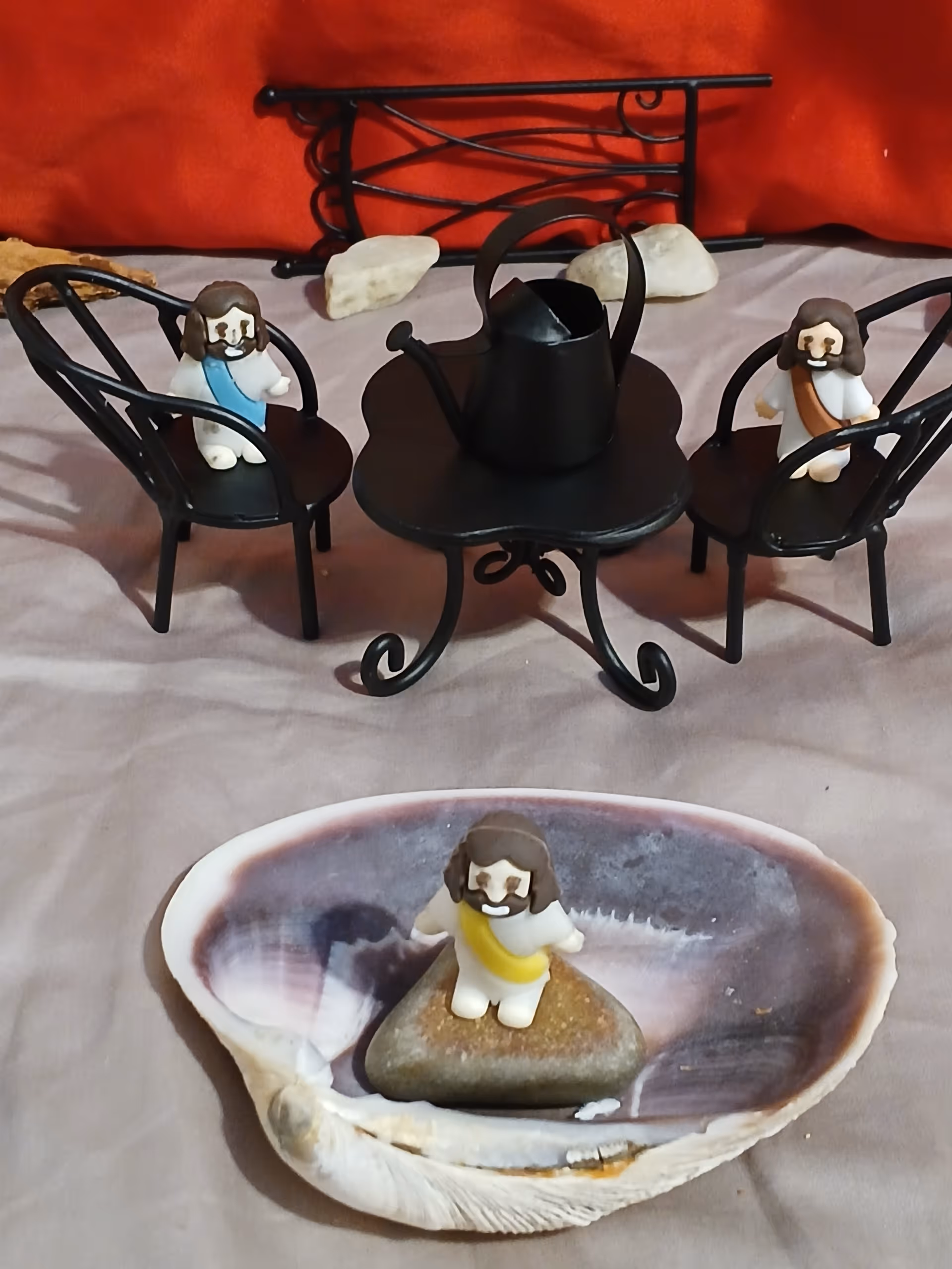 Small tabletop scene with miniature figurines seated around a black metal table and a figurine standing on a rock inside a large shell in the foreground.