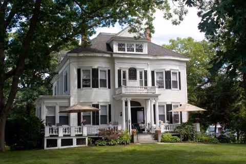 A large white residential building with a front porch, green lawn, and outdoor seating.