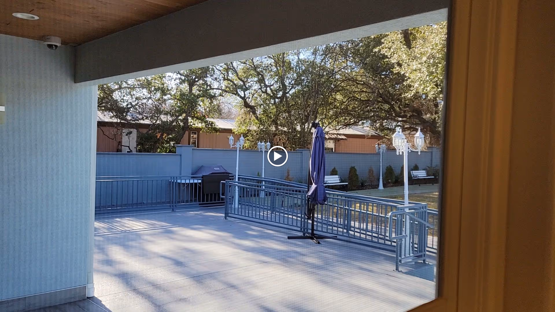 View of an outdoor patio area with a metal railing and a closed blue umbrella. Beyond the patio, there is a fenced yard with benches, lamp posts, and trees providing shade.