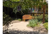 Sunlit courtyard with a curved brick path, a wooden bench under trees, surrounding shrubs, and a small hanging birdhouse.
