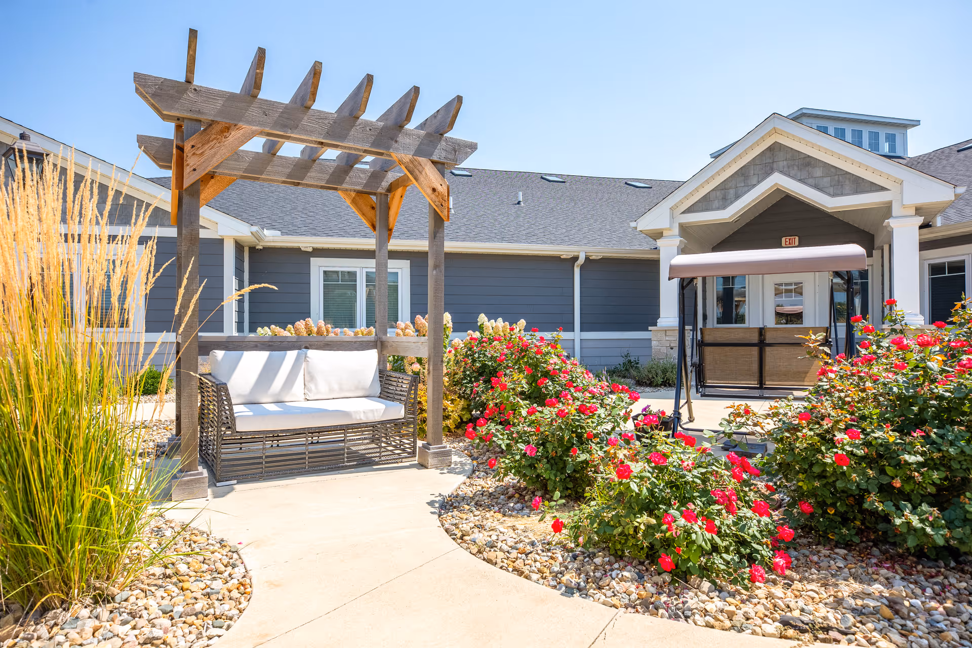 Outdoor seating area at Villas of Holly Brook Assisted Living & Memory Care in Streator, IL, featuring a wooden pergola with a cushioned bench swing, surrounded by landscaped garden beds with red flowers and ornamental grasses, and the facility building in the background under a clear blue sky.