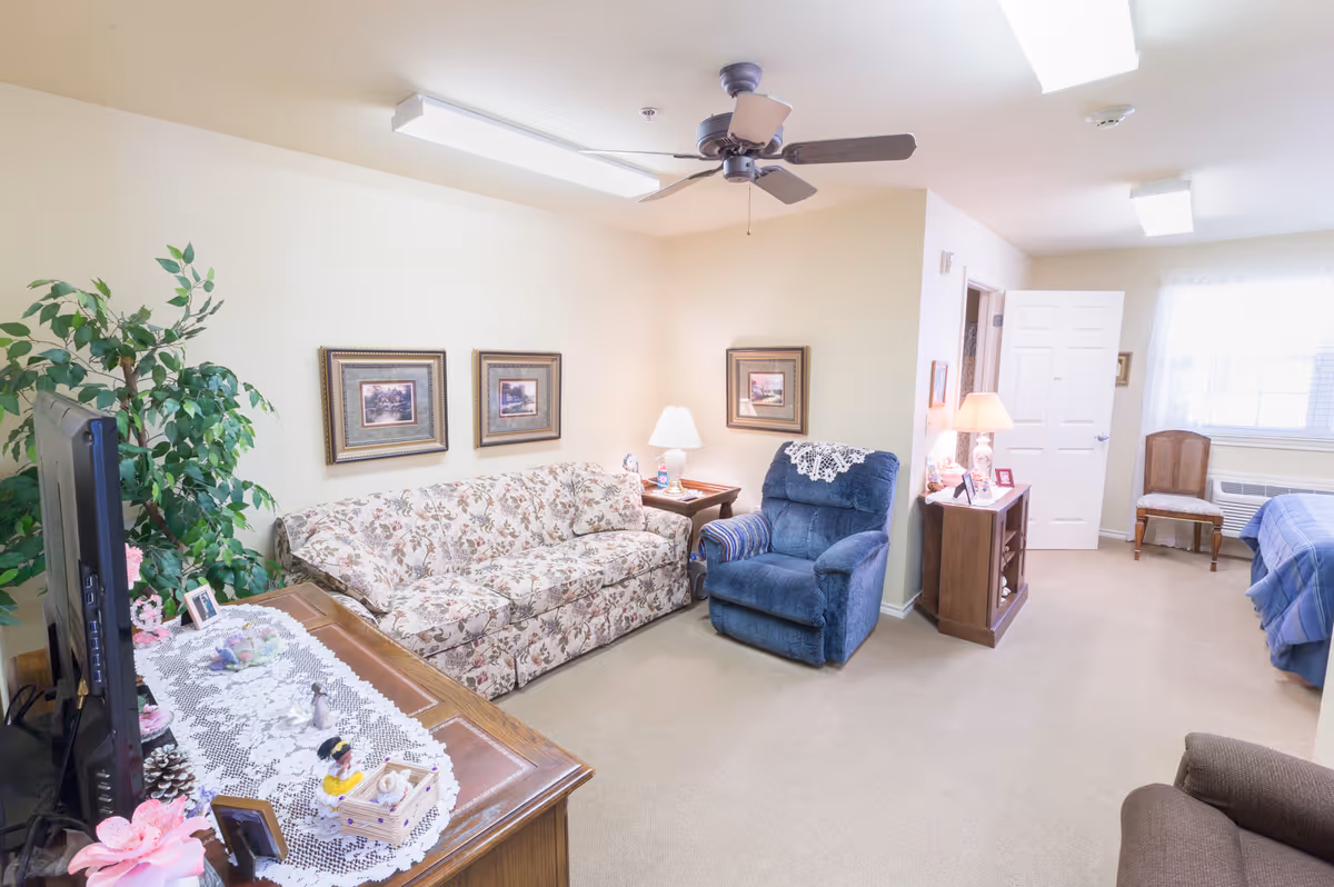 A cozy living area in a senior living facility featuring a floral-patterned sofa, a blue recliner with a lace doily on the back, a wooden TV stand with a lace runner and decorative items, framed artwork on the walls, a ceiling fan, and a small table with a lamp. In the background, there is a bedroom area with a bed covered in a blue blanket, a wooden chair, and a window with sheer curtains.