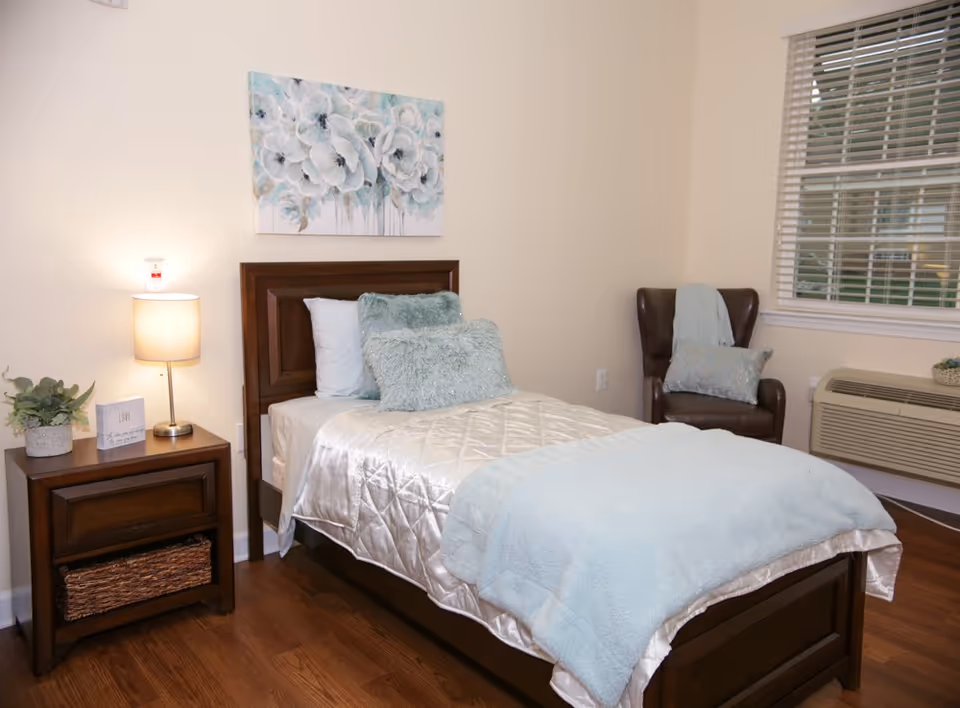 A neatly made single bed with white and light blue bedding in a bedroom. There is a wooden nightstand with a lamp, a small plant, and a decorative block with the word 'LOVE' on it. A painting of white flowers hangs above the bed. A brown armchair with a pillow and a light blue throw is placed near a window with white blinds. An air conditioning unit is installed below the window.