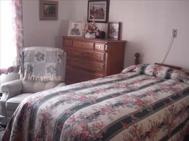 A bedroom with a bed covered in a floral patterned bedspread, a wooden dresser with framed photos and a flower arrangement on top, and a cushioned armchair with a patterned throw draped over the back. The room has light-colored walls and a window with floral curtains.
