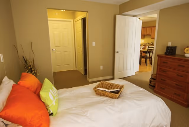 A bedroom with a bed featuring white bedding and colorful pillows in orange and green. A wicker basket with a book is placed on the bed. The room has beige walls and carpeted floor. There is a wooden dresser on the right side and an open door leading to another room with a dining table and chairs.