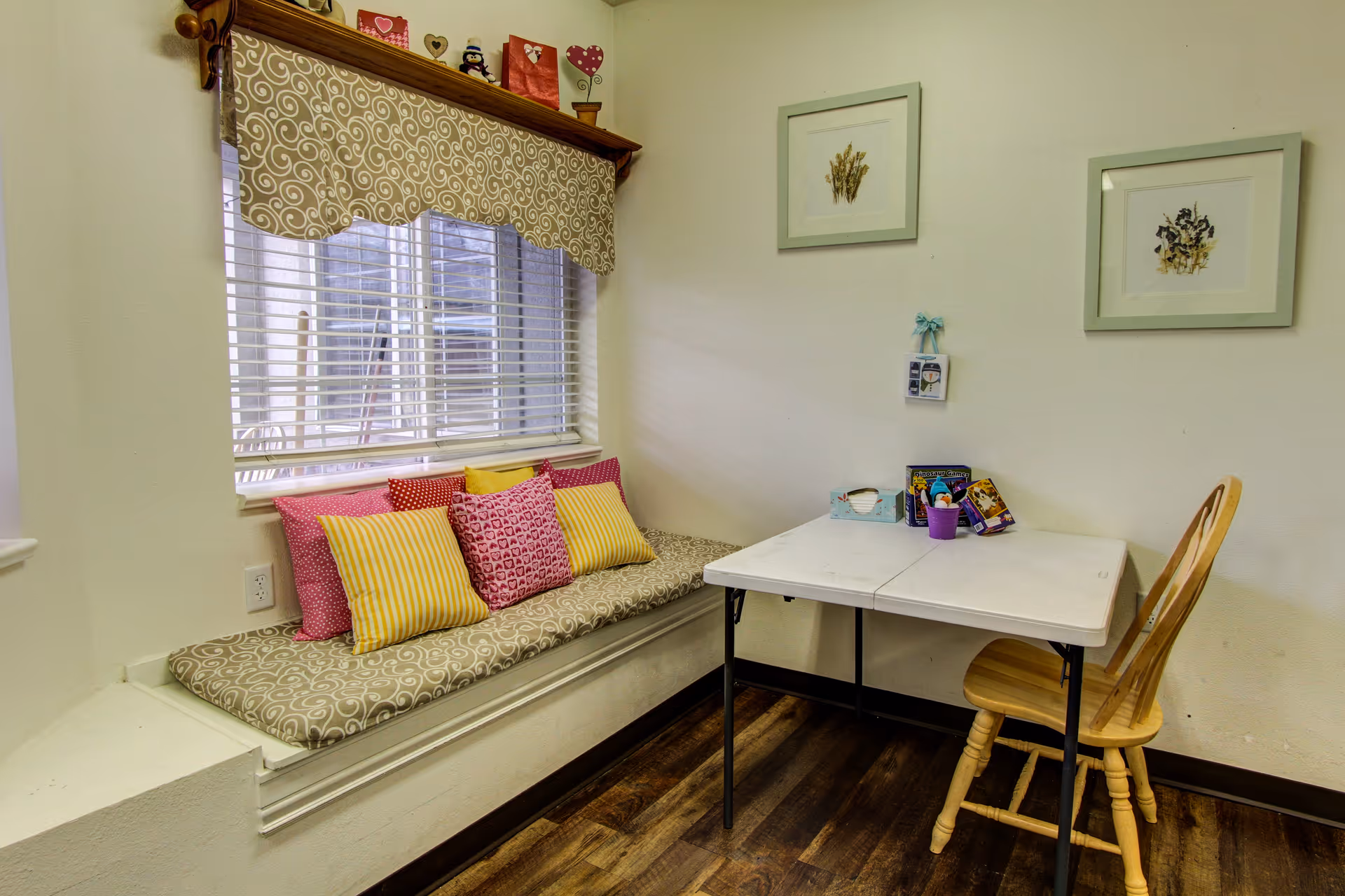 A cozy corner with a window seat featuring a patterned cushion and several colorful pillows in pink and yellow. Next to the window seat is a small white folding table with a wooden chair. On the table are some small items including a purple container and a tissue box. The walls are light-colored with two framed botanical prints hanging above the table.